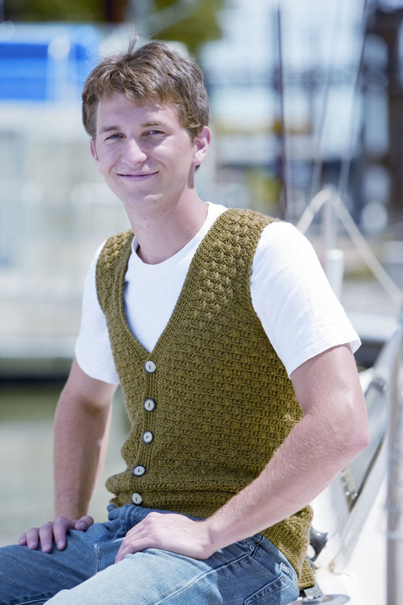 May include: A man wearing a brown knitted vest with buttons over a white t-shirt and blue jeans.