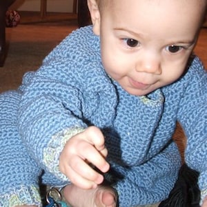 May include: A young child wearing a blue knitted sweater sits on the floor. The child is looking down at the camera. The sweater has a textured pattern and is a light blue color.