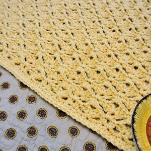 Pode incluir: Uma manta de crochê amarela com um padrão delicado e rendado. A manta está drapeada sobre uma superfície de tecido estampado.