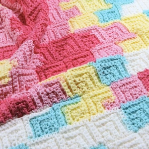 May include: A colourful crocheted blanket with a chevron pattern in shades of pink, yellow, blue and white.