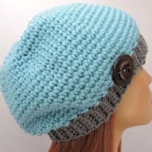 May include: A blue and gray knitted beanie with a large brown button. The beanie is made of a soft, textured yarn.