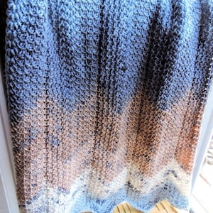 Op de afbeelding: Een blauwe, bruine en witte gebreide deken met een golvende rand. De deken is gemaakt met een textuur-steek en heeft een kleurverloop.