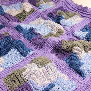 Puede incluir: Una manta de ganchillo con un esquema de color morado, azul y gris. La manta está hecha con diferentes puntos texturizados, creando un patrón único e interesante.