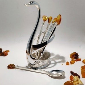 May include: Silver swan-shaped spoon holder with five spoons with amber handles. The holder is on a white surface with amber stones scattered around it.