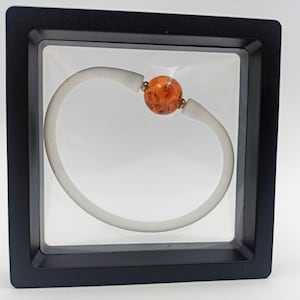 Op de afbeelding: Een witte rubberen armband met een enkele amberen kraal in het midden. De armband wordt getoond in een zwart vierkant frame met een transparante plastic insert.