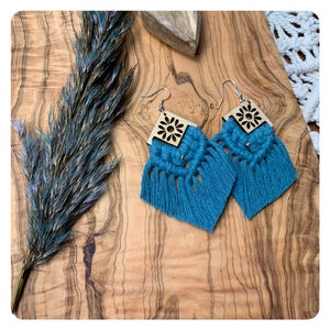 Könnte beinhalten: Türkise Quastenohrringe mit einem quadratischen Holzanhänger mit einem geschnitzten Blumendesign.