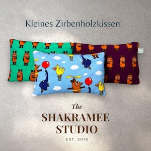 Könnte beinhalten: Drei dekorative Kissenbezüge mit verschiedenen Designs. Der erste Kissenbezug ist grün mit einem sich wiederholenden Muster aus braunen und orangefarbenen Comic-Tieren. Der zweite Kissenbezug ist hellblau mit einem sich wiederholenden Muster aus Comic-Tieren in einem Heißluftballon. Der dritte Kissenbezug ist lila mit einem sich wiederholenden Muster aus braunen und orangefarbenen Comic-Tieren. Der Text "Kleines Zirbenholzkissen" befindet sich oben im Bild. Der Text "The Shakramee Studio Est. 2018" befindet sich unten im Bild.