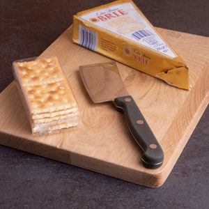 Può includere: Un tagliere in legno con una porzione di formaggio Brie, un piccolo coltello e un pacchetto di cracker. Il Brie è in un involucro di carta dorata. Il coltello ha un manico nero. I cracker sono in un involucro di plastica trasparente.
