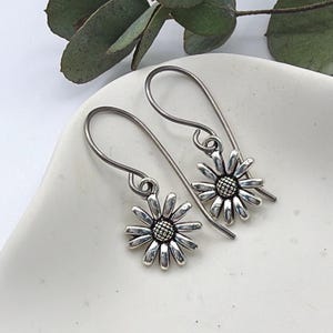 May include: A pair of silver daisy earrings. Each earring features a detailed daisy flower with a dark centre and delicate petals. The earrings have a hook design for pierced ears, and are displayed on a white surface.