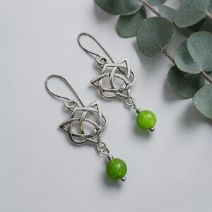 May include: Silver-toned Celtic knot earrings with green gemstone beads. The earrings have a star-shaped knot design with a small, round, bright green bead dangling below. The earrings are displayed on a white surface.