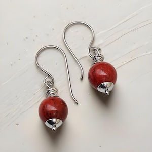 May include: A pair of red coral bead earrings with silver-colored metal accents. The earrings feature a round, textured coral bead suspended from a silver hook. The beads are approximately 0.5 inches in diameter.