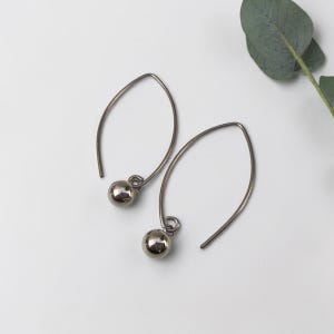 May include: A pair of silver-colored, teardrop-shaped earrings. Each earring features a curved wire design with a small, round, metallic bead dangling at the bottom. The earrings are displayed on a white surface, with a green leaf in the background.