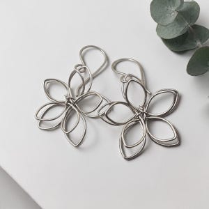 May include: A pair of silver flower earrings. Each earring has an openwork design with five petals. The earrings hang from simple ear wires. The background is a clean white surface with a sprig of green leaves.