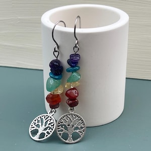 May include: A pair of dangle earrings featuring a silver tree of life charm and a colorful stack of gemstones. The gemstones include amethyst, blue, green, yellow, red, and brown. The earrings are displayed next to a white ceramic container.
