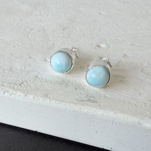 May include: A pair of stud earrings featuring round, polished light blue Larimar gemstones set in simple silver bezels. The stones exhibit a smooth, opaque surface with subtle white marbling patterns, securely mounted on silver posts.