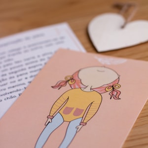 May include: A pink card with a cartoon illustration of a girl with pink hair wearing a yellow shirt with a pink pocket and blue pants. The girl is smiling and has her eyes closed.