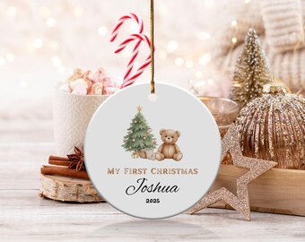 Puede incluir: Un adorno navideño de cerámica blanca con una cuerda dorada para colgar. El adorno presenta una ilustración de acuarela de un árbol de Navidad y un oso de peluche. El texto dice "MY FIRST CHRISTMAS" y "Joshua 2025".