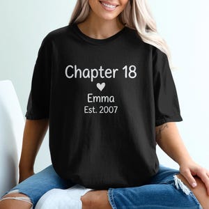 May include: Black t-shirt with white text that reads "Chapter 18" above a heart, the name "Emma", and the year "Est. 2007". The shirt is worn by a person sitting on a white chair.