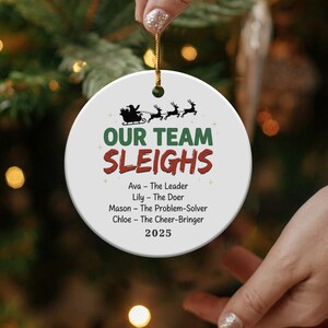 May include: A white, round Christmas ornament with the text "OUR TEAM SLEIGHS" in green and red. The ornament features a silhouette of Santa's sleigh and reindeer, with names and roles below, and the year 2025.