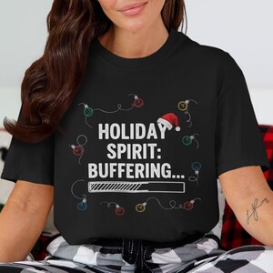 Puede incluir: Camiseta negra con el texto blanco "HOLIDAY SPIRIT: BUFFERING...", con un gorro de Papá Noel y luces de cadena. La camiseta tiene un gráfico de barra de carga, lo que sugiere una visión humorística de la anticipación navideña.