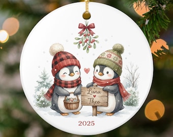 May include: A round Christmas ornament featuring two penguins wearing hats and scarves, holding a sign that reads "Luna Theo" with a heart symbol. The ornament has a 2025 date and a mistletoe design at the top.