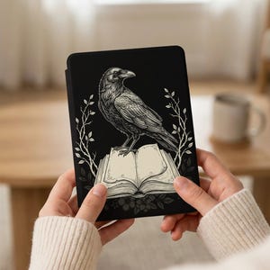 May include: Black tablet case featuring a detailed illustration of a raven perched atop an open book. The raven is rendered in grayscale, with intricate feather details. White floral and branch accents frame the design against a black background.