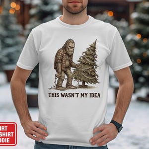 May include: White t-shirt featuring a Bigfoot character struggling with a Christmas tree and string lights. The text "THIS WASN'T MY IDEA" is printed below the graphic. The shirt is a casual wear item.