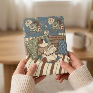 May include: A rectangular case featuring a cat reading a book while lounging on a striped cushion. The case has a blue floral pattern on the top and a striped pattern on the bottom. The cat is wearing a sweater.