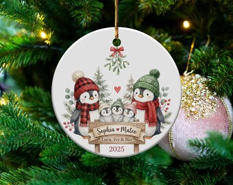May include: A round Christmas ornament featuring a family of penguins wearing hats and scarves. The ornament has a wooden sign with the names "Sophia & Mateo" and "Luca, Ivy & Rio" and the year 2025. The background includes trees and a heart.