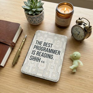 Könnte beinhalten: Ein Notizbuch mit dem Text "THE BEST PROGRAMMER IS READING. SHHH 👀" liegt auf einer Holzoberfläche, neben einer Sukkulente in einem gemusterten Topf, einer brennenden Kerze, einem kleinen Wecker und einer kleinen Figur.