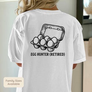 May include: White t-shirt featuring a black illustration of an open egg carton with eggs. The text "EGG HUNTER (RETIRED)" is printed below the graphic. The shirt also has a small label that says "Family Sizes Available".