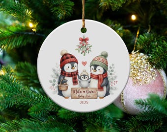 May include: A round Christmas ornament featuring a penguin family. The penguins wear winter hats and scarves. The ornament has the text "Mila & Owen Baby Arlo 2025". A red bow and mistletoe are at the top.