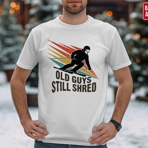 May include: White t-shirt featuring a skier graphic with the text "OLD GUYS STILL SHRED" in black. The skier is depicted in black, with a colorful retro design. A red box in the top right corner says "BACK PRINT AVAILABLE".