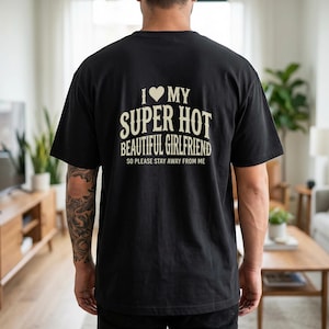 May include: Black t-shirt with the text "I ❤️ MY SUPER HOT BEAUTIFUL GIRLFRIEND SO PLEASE STAY AWAY FROM ME" in a cream-colored font. The shirt is a casual style, likely made of cotton or a similar fabric.