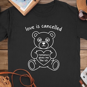 Puede incluir: Camiseta negra con un dibujo blanco de un oso de peluche sosteniendo un corazón. El texto "love is cancelled" está encima del oso, y "romance gives me the ick" está en el corazón. La camiseta se muestra sobre una superficie de madera.