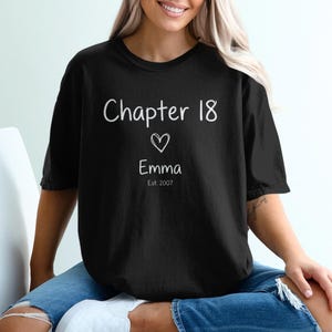 Peut inclure: T-shirt noir avec l'inscription blanche "Chapter 18", un cœur, le prénom "Emma" et "Est. 2007". La personne porte le t-shirt et est assise sur une chaise blanche, vêtue d'un jean.