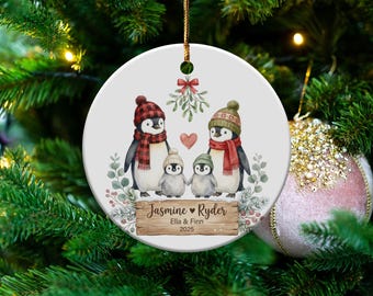 May include: A round Christmas ornament featuring a family of penguins wearing hats and scarves. The ornament has a wooden sign with the names "Jasmine & Ryder" and the year "2025". A red heart and mistletoe are also featured.
