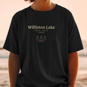 Puede incluir: Camiseta negra con el texto "Williston Lake" y las coordenadas 56.0° N -124.5° W, Canadá. Debajo, un gráfico de un lago con árboles y una canoa. La camiseta es de estilo casual.