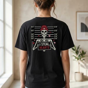 May include: Black t-shirt with a skeleton design wearing a red beanie and holding a sign saying "I STEAL HEARTS." The design includes a height chart with measurements in feet and inches, and pink hearts.