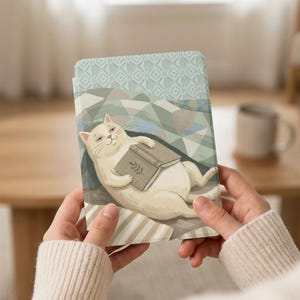 May include: A tablet case featuring a whimsical illustration of a relaxed white cat holding a book. The case has a light blue and green geometric pattern on the top and a striped pattern on the bottom. A mug is in the background.