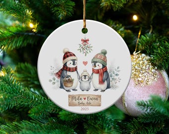 May include: A round Christmas ornament featuring a family of penguins wearing hats and scarves. The ornament has the text "Mila Owen Baby Arlo 2025" on a wooden sign. The ornament is hanging on a Christmas tree.
