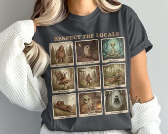 May include: Dark gray t-shirt featuring a grid of illustrated panels with cryptids and aliens. The text "Respect the Locals" is printed across the top. Each panel depicts a different scene with text such as "Space Appreciated" and "It's Okay to Hide."