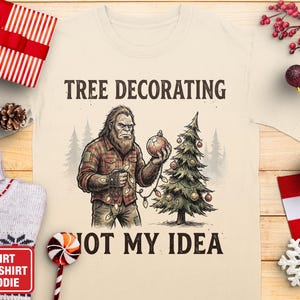 May include: Cream-colored t-shirt with a graphic of Bigfoot holding a Christmas ornament and string lights, next to a decorated Christmas tree. Text reads "TREE DECORATING NOT MY IDEA". Festive holiday apparel.