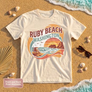 Puede incluir: Camiseta color crema con un gráfico de Ruby Beach, Washington, con una puesta de sol, olas del océano y una torre de salvavidas. El diseño incluye el texto "Ruby Beach Washington". Gafas de sol y conchas marinas sobre la arena.