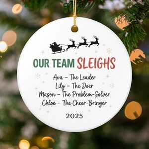 May include: White ceramic Christmas ornament with a gold string. The ornament features a black silhouette of Santa's sleigh and reindeer, with the text "OUR TEAM SLEIGHS" in green and red. Below are names and titles, and the year 2025.