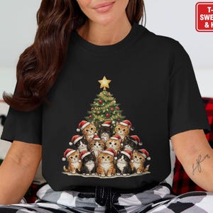 May include: Black t-shirt featuring a Christmas tree made of kittens wearing Santa hats. The tree is topped with a gold star. The shirt has the text "T-SHIRT SWEATSHIRT & HOODIE" in a red box.