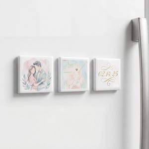 May include: Three white square magnets with watercolor designs. One features a couple embracing, another a map with a heart, and the last displays the date "02.14.25" in gold script. The magnets are attached to a white surface.