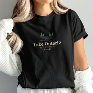 Op de afbeelding: Zwart T-shirt met een afbeelding van Lake Ontario, inclusief bomen, water en een kano. De tekst "Lake Ontario" staat boven de coördinaten 43,7° N -77,9° W, en "Canada" eronder. Een casual kledingstuk.