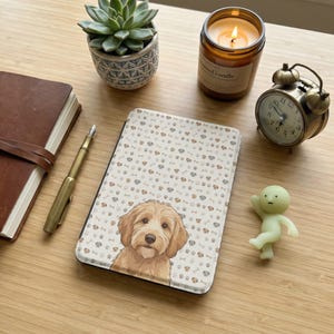 Puede incluir: Una funda de tableta beige y blanca con una ilustración de Labradoodle. Rodeada por un diario de cuero marrón, una planta suculenta, una vela, un despertador vintage, un bolígrafo y una pequeña figura.