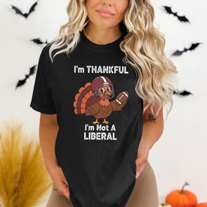Könnte beinhalten: Schwarzes T-Shirt mit einem Thanksgiving-Motiv. Das Design zeigt einen Cartoon-Truthahn mit Footballhelm und Football. Der Text auf dem Shirt lautet "I'm THANKFUL" über "I'm Not A LIBERAL".